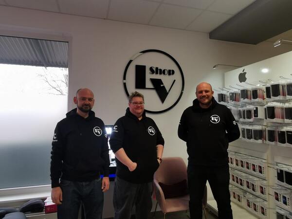 Drei Mitarbeiter in schwarzer IT-Shop Kleidung stehen im Handyshop vor Zubehörwand und Logo.
