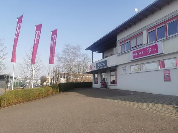 Telekom Shop mit Firmengebäude, Partner-Logo und Fahnen auf dem Parkplatz bei Sonnenschein.