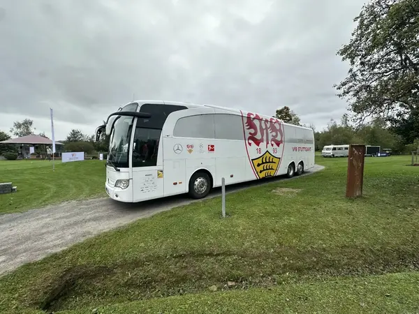 Ein weißer Mannschaftsbus mit VfB Stuttgart Logo steht auf einer grünen Wiese im Freien.