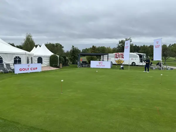 Golfplatz mit weißen Zelten, Golf Cup Bannern und VfB Stuttgart Bus bei bewölktem Himmel.